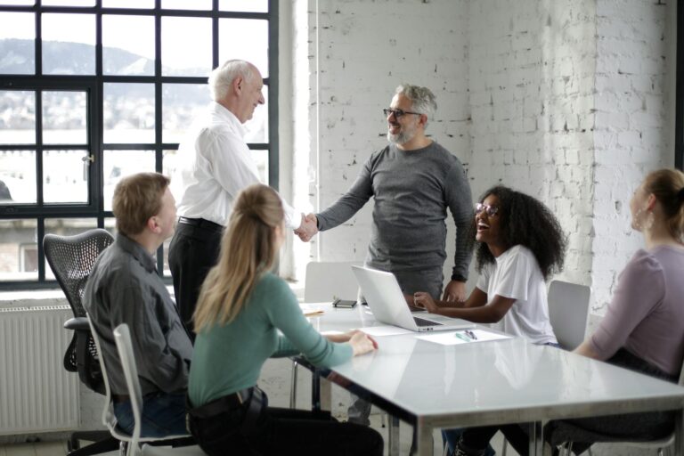 Cheerful male colleagues shaking hands while discussing business ideas with group of multiethnic coworkers gathering around table with gadgets and documents in modern light workspace